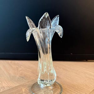 Pode incluir: Um vaso de vidro transparente com um design semelhante a uma flor. O vaso tem um topo alargado com múltiplas aberturas em forma de pétala e uma haste fina e torcida. Assenta numa base circular, mostrando uma estética delicada e elegante.