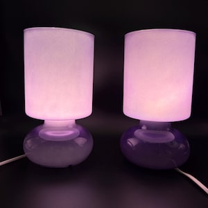 May include: Two table lamps with cylindrical frosted glass shades that emit a soft purple glow. Each lamp features a rounded, translucent purple glass base and a white power cord extending from the bottom, set against a dark background.