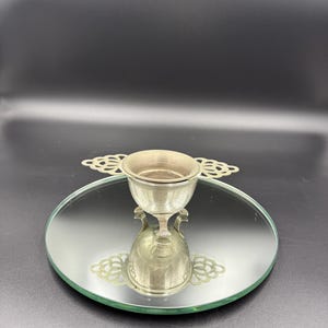 May include: A silver-colored, ornate cup with a decorative handle, resting on a round, mirrored surface. The cup has a small bowl and a detailed base. The mirrored surface reflects the cup, creating a symmetrical image. The background is a solid black.