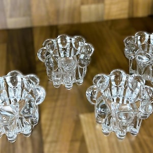 May include: Four clear glass candle holders with a bubble-like design. The candle holders are arranged on a wooden surface. The glass is transparent, allowing light to pass through, creating a decorative effect.
