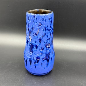 Könnte beinhalten: Eine blaue Keramikvase mit strukturierter, getropfter Glasur. Die Vase hat eine zylindrische Form mit leicht ausgestelltem Boden und dunklem braunen Innenraum. Die Außenseite weist Blautöne auf.