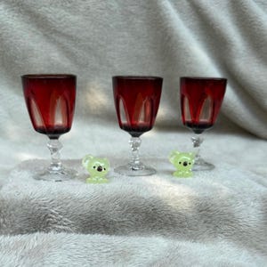 Può includere: Tre calici di vino rosso rubino con steli trasparenti sono disposti su una superficie bianca e morbida. Due piccole statuette di koala verdi sono poste vicino ai calici. I calici hanno un design scanalato unico.