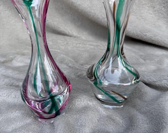 May include: Two clear glass vases with a flared top and a slender body. One vase has pink and green stripes, the other has green and brown stripes. The vases are decorative and suitable for displaying flowers.