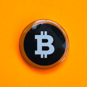 Könnte beinhalten: Ein runder Button mit einem schwarzen Zentrum, das ein weißes Bitcoin-Symbol zeigt. Der Button hat einen braunen Rand und ist vor einem leuchtend orangefarbenen Hintergrund platziert. Das Design ist schlicht und modern.