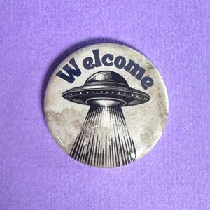 Könnte beinhalten: Ein runder Button mit einer Illustration im Vintage-Stil einer fliegenden Untertasse, die einen Lichtstrahl aussendet. Das Wort "Welcome" ist in dunkler blauer Schrift über dem Raumschiff angeordnet. Der Hintergrund ist verwaschen grau.