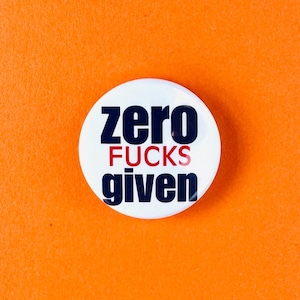May include: A white button with the text "zero FUCKS given" in navy blue and red lettering. The button is centered on an orange background.