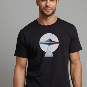 May include: Black t-shirt with a graphic of a UFO above a figure. The UFO is blue, red, and white, set against a white circle. The figure is descending from the UFO. The shirt is made of a soft material.