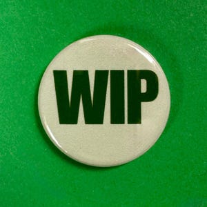 Op de afbeelding: Een ronde, witte button met de letters "WIP" in een vetgedrukte, donkergroene lettertype. De button is gecentreerd op een effen groene achtergrond. De button heeft een glanzende afwerking en een licht getextureerd oppervlak.