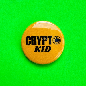 Könnte beinhalten: Ein runder, gelber Button mit dem schwarzen Schriftzug "CRYPT KID". Rechts neben dem Wort "CRYPT" befindet sich eine kleine Grafik eines "C" in einem Kreis. Der Button ist vor einem leuchtend grünen Hintergrund platziert.