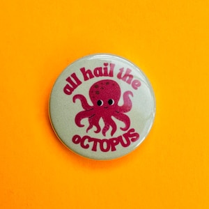 May include: A circular badge with the words "all hail the octopus" in red. The badge features a red cartoon octopus with a smiling face. The badge has a light green background, set against an orange backdrop.
