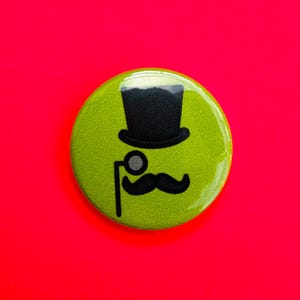 Puede incluir: Un botón redondo verde lima con un diseño de sombrero de copa negro, monóculo y bigote. El botón está sobre un fondo rojo brillante. El diseño es una versión caprichosa de los accesorios de un caballero.