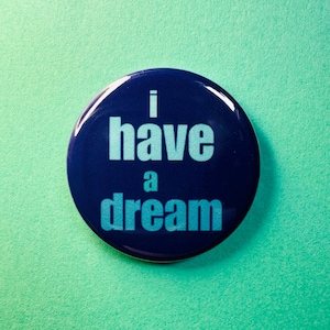 Puede incluir: Un botón redondo azul marino con la frase "i have a dream" en azul claro. El botón está sobre un fondo verde claro. El diseño es simple y audaz.
