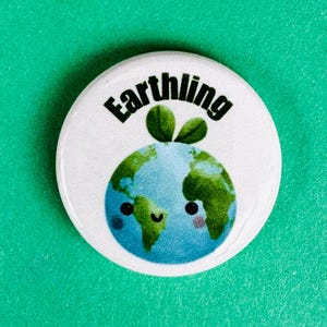 May include: A circular badge with the word "Earthling" in black, curved text. The badge shows a cartoon globe with blue oceans, green continents, and a smiling face. Two green leaves are at the top of the globe.