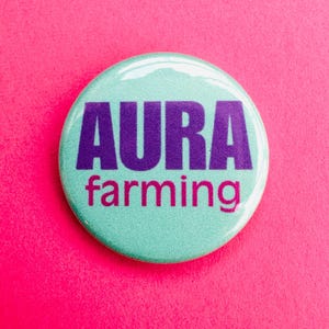 Könnte beinhalten: Ein runder, türkisfarbener Button mit den lila Worten "AURA" und den pinkfarbenen Worten "farming". Der Button befindet sich vor einem hellrosa Hintergrund.