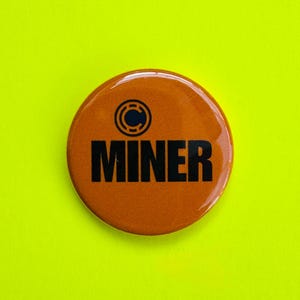 Könnte beinhalten: Ein runder, orangefarbener Button mit dem Wort "MINER" in fetten, schwarzen Buchstaben. Oberhalb des Wortes befindet sich ein kreisförmiges Design mit blauem Zentrum. Der Button befindet sich vor einem hellgelb-grünen Hintergrund.