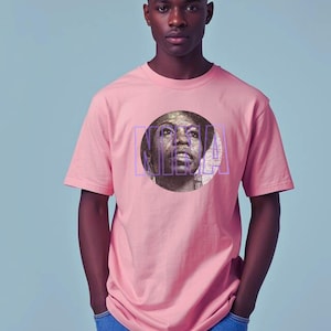 May include: A light pink t-shirt featuring a portrait of a woman within a purple rectangular frame. The person in the image is wearing the t-shirt and blue jeans. The background is a solid light blue.