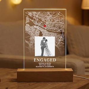 May include: A personalized engagement gift featuring a map design with a red heart, a photo, and the word "ENGAGED". The wooden base supports a clear acrylic panel with the couple's names and date.