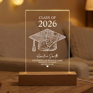 Op de afbeelding: Een helder acryl afstudeercadeau met een houten voet. Het ontwerp bevat de tekst "CLASS OF 2026", een afstudeerhoed met een kaartontwerp, de naam "Amelia Smith" en andere details. De lamp is verlicht.