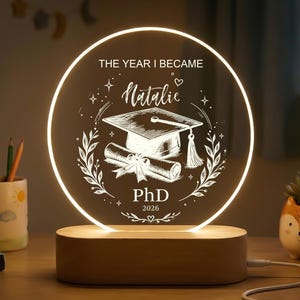 May include: A graduation-themed LED lamp with a circular illuminated design. The lamp displays the text "THE YEAR I BECAME Natalie PhD 2026" with a graduation cap and scroll. The lamp is set on a wooden base.