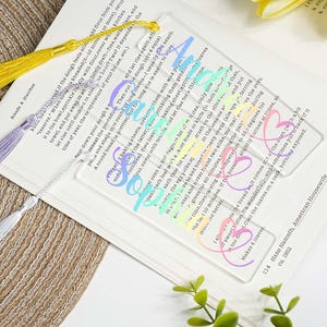 May include: Clear acrylic bookmarks with colorful text that reads "Soulmate" and a heart design. Each bookmark has a tassel in yellow, lavender, or silver. The bookmarks are on a book page.