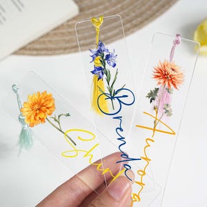 May include: Three clear acrylic bookmarks with floral designs and names. One features an orange flower, another blue flowers, and the third an orange and pink flower. Each has a tassel and a name written in a script font.