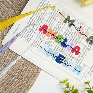 Peut inclure: Marque-pages en acrylique transparents avec le nom "amelia" en lettres colorées et des pompons assortis. Les marque-pages sont sur une page de livre. Les pompons sont jaunes, violets et argentés.