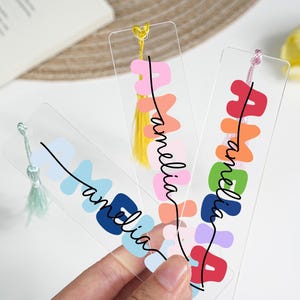 May include: Three clear acrylic bookmarks with colorful designs and the name "amelia" in a cursive font. Each bookmark has a different color tassel. The background includes a book and a decorative mat.