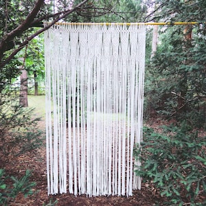 May include: A white macrame wall hanging suspended from a wooden dowel, set against a backdrop of lush green trees and foliage. The intricate woven design creates a textured, bohemian aesthetic, ideal for home decor.