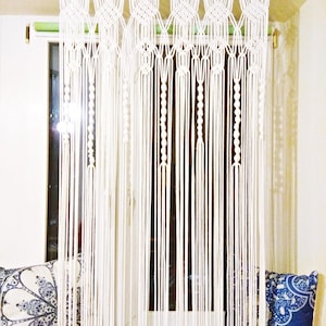 Op de afbeelding: Witte macramé gordijn dat voor een raam hangt. Het gordijn heeft een geometrisch ontwerp aan de bovenkant en lange, verticale strengen. Decoratieve kussens in blauw en wit zijn zichtbaar op de vensterbank.