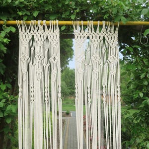 Op de afbeelding: Crèmekleurige macramé gordijnen hangend aan een gele metalen stang, omlijst door weelderig groen gebladerte. De gordijnen hebben ingewikkelde knoop patronen en lange, verticale strengen, waardoor een textuurrijke, bohemien esthetiek ontstaat. Het natuurlijke licht filtert door de open ruimte.