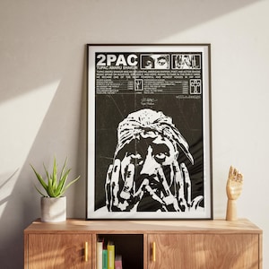 May include: Black and white framed poster featuring a portrait of Tupac Shakur. The poster includes text with biographical information and album titles. The image is displayed on a wooden cabinet with a plant and a wooden hand sculpture.