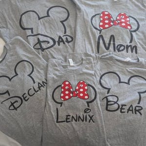 May include: Five gray short-sleeved t-shirts featuring black outlines of mouse ears. Two shirts display red and white polka-dot bows. The shirts are printed with the text Dad, Mom, Declan, Lennix, and Bear in a stylized black script font.