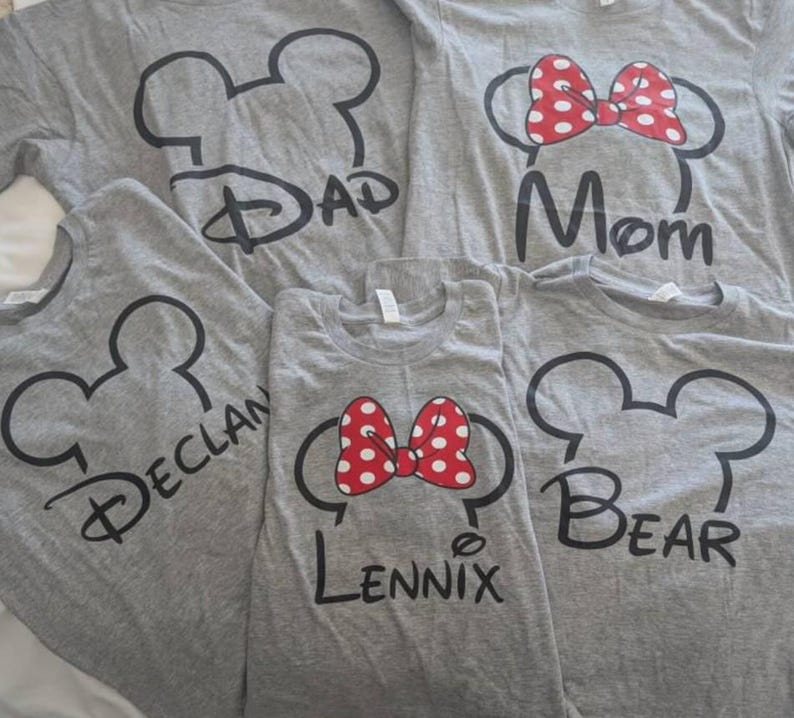 May include: Five gray short-sleeved t-shirts featuring black outlines of mouse ears. Two shirts display red and white polka-dot bows. The shirts are printed with the text Dad, Mom, Declan, Lennix, and Bear in a stylized black script font.