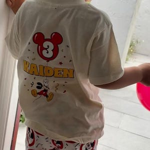 May include: A cream-colored short-sleeve t-shirt featuring a graphic print on the back. The design includes a red Mickey Mouse-shaped balloon with the number 3 inside, the name KAIDEN in yellow letters, and a small Mickey Mouse character below.