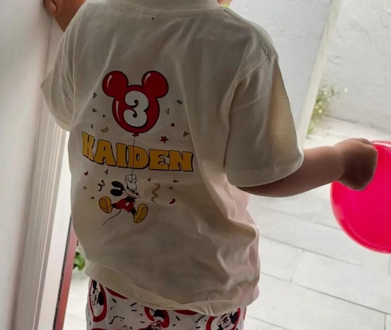 May include: A cream-colored short-sleeve t-shirt featuring a graphic print on the back. The design includes a red Mickey Mouse-shaped balloon with the number 3 inside, the name KAIDEN in yellow letters, and a small Mickey Mouse character below.