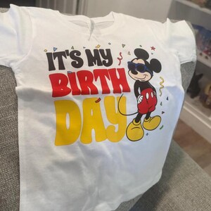 May include: A white short-sleeved t-shirt featuring a graphic of Mickey Mouse wearing blue sunglasses. The text reads IT'S MY BIRTH DAY in black, red, and yellow lettering. Small colorful confetti shapes are scattered around the design.