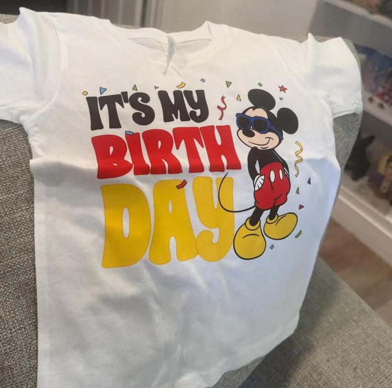 May include: A white short-sleeved t-shirt featuring a graphic of Mickey Mouse wearing blue sunglasses. The text reads IT'S MY BIRTH DAY in black, red, and yellow lettering. Small colorful confetti shapes are scattered around the design.