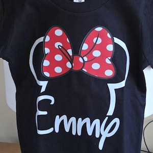 May include: A black short-sleeved t-shirt featuring a white outline of mouse ears topped with a red bow that has white polka dots. Below the ears, the name Emmy is printed in a white script font, centered on the front of the garment.