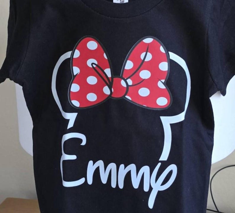 May include: A black short-sleeved t-shirt featuring a white outline of mouse ears topped with a red bow that has white polka dots. Below the ears, the name Emmy is printed in a white script font, centered on the front of the garment.