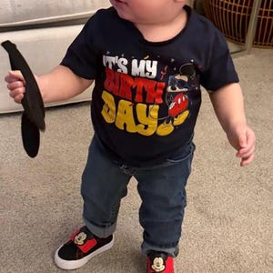 May include: A navy blue short-sleeve t-shirt featuring the text IT'S MY BIRTHDAY in white, red, and yellow, accompanied by a graphic of a cartoon mouse wearing sunglasses. The outfit includes dark blue denim jeans and black and red sneakers.