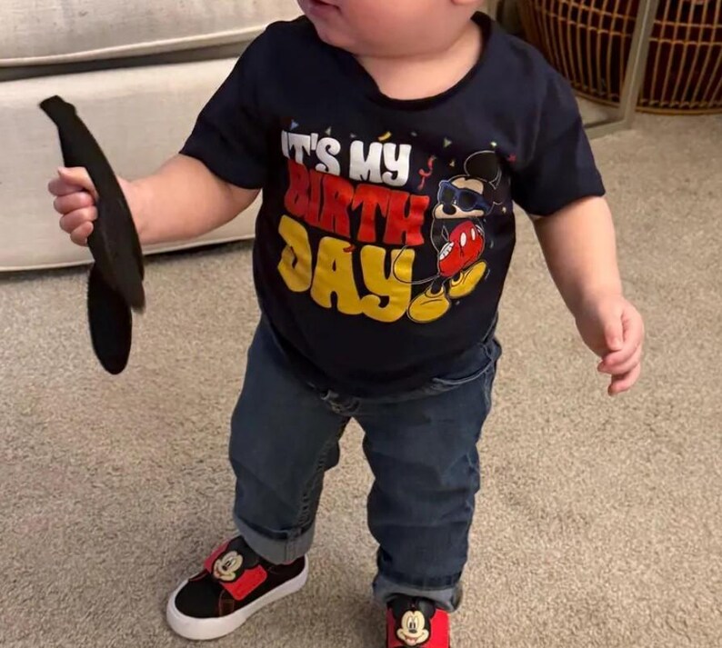 May include: A navy blue short-sleeve t-shirt featuring the text IT'S MY BIRTHDAY in white, red, and yellow, accompanied by a graphic of a cartoon mouse wearing sunglasses. The outfit includes dark blue denim jeans and black and red sneakers.