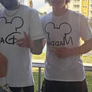 May include: Two white short-sleeved t-shirts featuring a black graphic of a mouse ear silhouette. The left shirt displays the word DAD in a stylized font, while the right shirt features the word MADDEN written in a similar black script font.