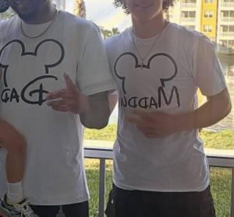 May include: Two white short-sleeved t-shirts featuring a black graphic of a mouse ear silhouette. The left shirt displays the word DAD in a stylized font, while the right shirt features the word MADDEN written in a similar black script font.