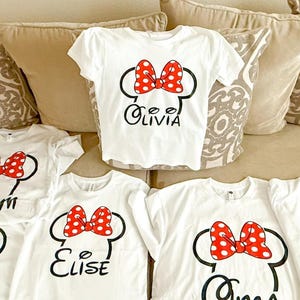 May include: A collection of white short-sleeved t-shirts featuring a black outline of mouse ears with a red and white polka-dot bow. Each shirt displays a unique name in black script lettering, including the names Olivia, Elise, and others.