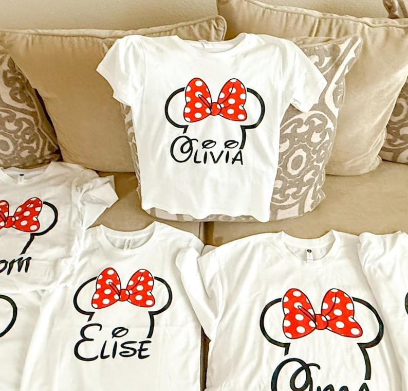 May include: A collection of white short-sleeved t-shirts featuring a black outline of mouse ears with a red and white polka-dot bow. Each shirt displays a unique name in black script lettering, including the names Olivia, Elise, and others.