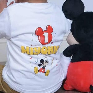 May include: A white short-sleeved t-shirt featuring a graphic print of a red Mickey Mouse-shaped balloon with the number 1 inside, the name MIYONNI in yellow letters, and a small Mickey Mouse character surrounded by colorful confetti.