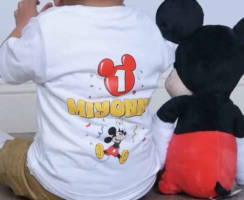 May include: A white short-sleeved t-shirt featuring a graphic print of a red Mickey Mouse-shaped balloon with the number 1 inside, the name MIYONNI in yellow letters, and a small Mickey Mouse character surrounded by colorful confetti.