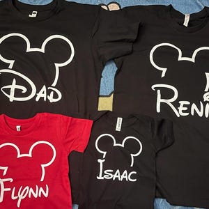 May include: Three black t-shirts and one red t-shirt, each featuring a white outline of a mouse head silhouette above a name printed in a stylized font. The names displayed are Dad, Reni, Flynn, and Isaac, all in a white graphic design.