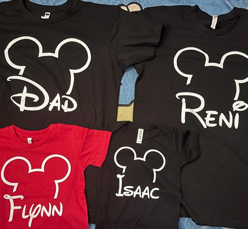 May include: Three black t-shirts and one red t-shirt, each featuring a white outline of a mouse head silhouette above a name printed in a stylized font. The names displayed are Dad, Reni, Flynn, and Isaac, all in a white graphic design.