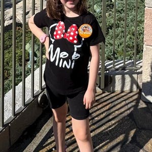 May include: A black short-sleeved t-shirt featuring a white outline of a bow and the text "MINI" in white. A red bow with white polka dots is positioned above the text. A round yellow "1st Visit" button is pinned to the upper chest area.